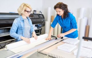 Two women stand at a wide format printer aligning the paper and assessing the output.