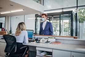 A professional male wearing a mask stands over a woman professional sitting at her desk working.