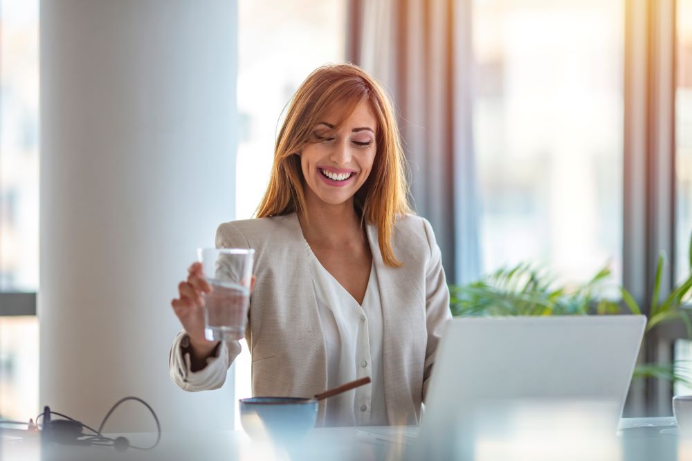 A businesswoman grabs a glass of water while sitting at a desk behind a computer signifying business office solutions.
