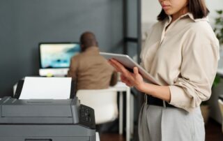 A young professional woman stands in the foreground next to an all in one office printer in a Charlotte office while a coworker diligently works in the background.