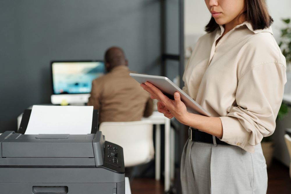 A young professional woman stands in the foreground next to an all in one office printer in a Charlotte office while a coworker diligently works in the background.