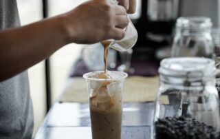 Close-up of a person pouring milk into a tall glass of iced coffee, prepared using ice from a commercial ice maker, on a cafe counter.
