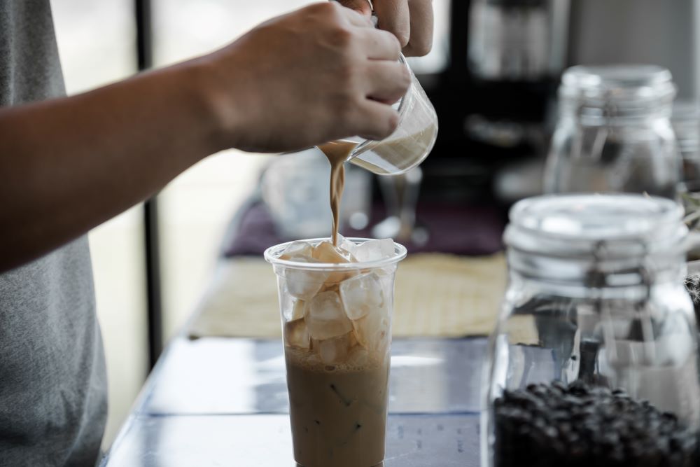 Close-up of a person pouring milk into a tall glass of iced coffee, prepared using ice from a commercial ice maker, on a cafe counter.