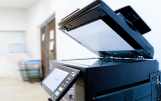 The image features a close-up of an all-in-one office printer with its scanning lid open, indicating its multifunctional capabilities. The printer has a modern design with a large, user-friendly interface displaying icons for various functions such as copy, scan, and fax. The focus on the printer's control panel and the open scanner bed emphasizes the machine's convenience for daily office tasks. Situated in a bright office environment with soft, natural light and blurred background elements, the printer stands ready for use, symbolizing the efficiency and productivity that such devices bring to the workplace.