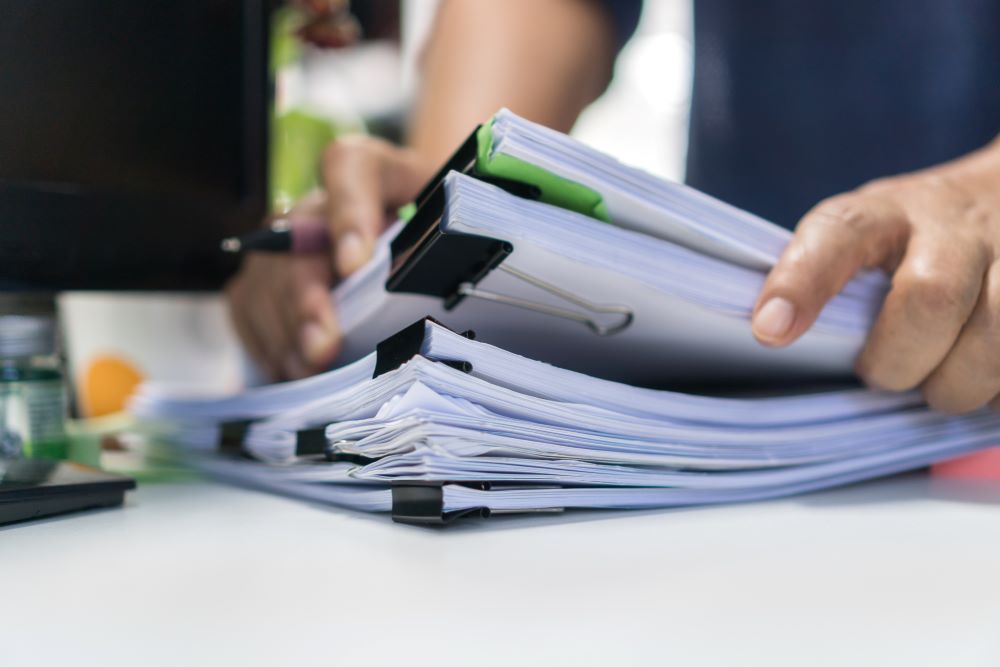 Close-up of a person's hands organizing a large stack of papers clipped into binders, demonstrating effective document management. The paperwork is on a white desk, with a computer monitor in the soft-focused background, highlighting an organized approach to handling office documents.