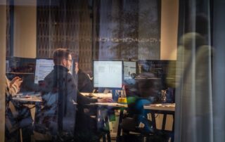 Office workers collaborating at their desks with multiple monitors and software tools visible, showcasing modern office IT solutions in action through a glass window.